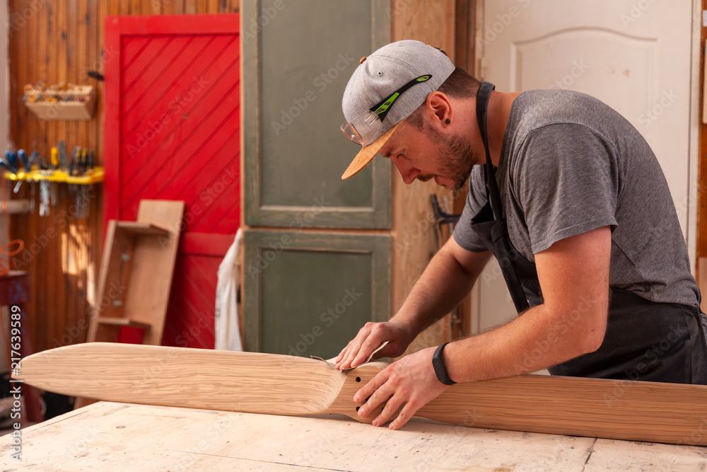 A man carpenter in a cap, goggles and a T-shirt is sanding a wooden in the workshop, in the background a lot of tools A man carpenter in a cap, goggles and a T-shirt is sanding a wooden in the workshop, in the background a lot of tools
