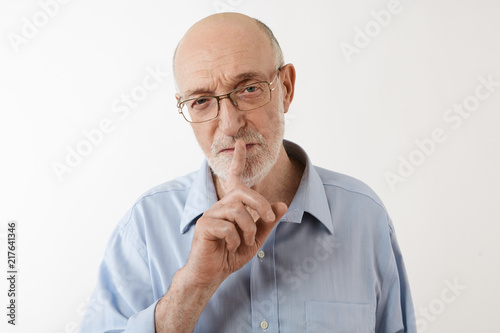 Attractive stylish elderly male wearing formal blue shirt and rectangular eyeglasses doing hush gesture, holding fore finger at his lips, asking to keep his secret. Confidential information