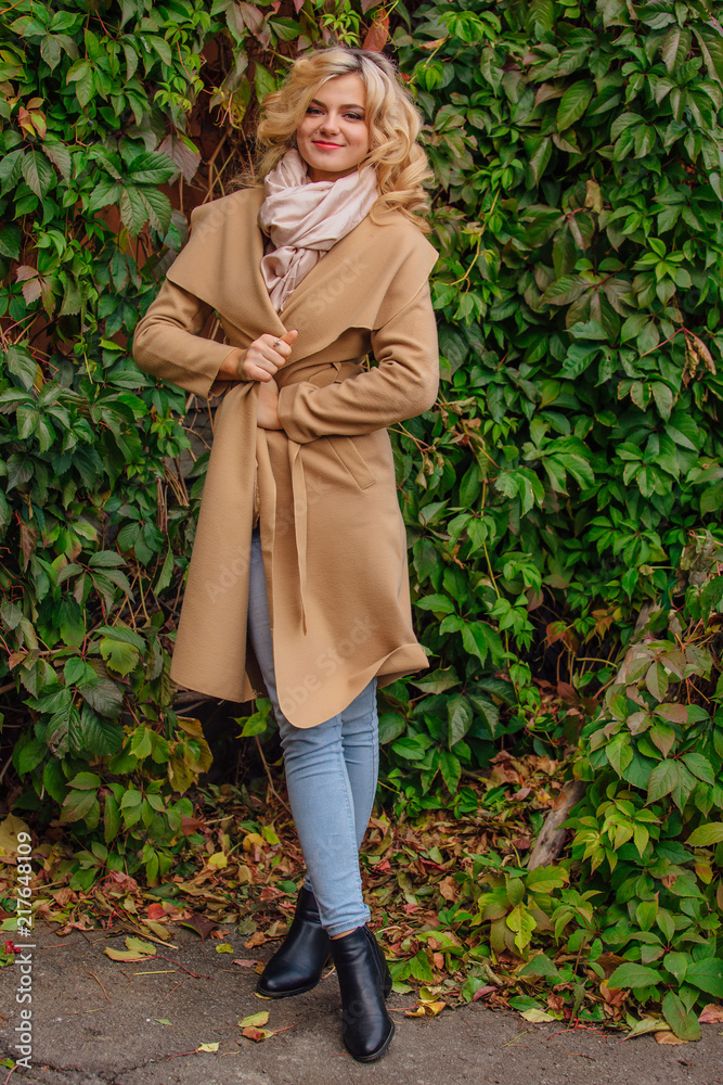 Yuong beautiful woman in sweater and coat stands next to the background of wild grapes leaves Yuong beautiful woman in sweater and coat stands next to the background of wild grapes leaves