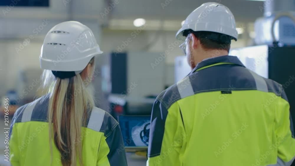 Following Shot of Male and Female Mechanical Engineers Talking, while Walking Through Industrial Factory, Male Holds Laptop With Engine Project. Following Shot of Male and Female Mechanical Engineers Talking, while Walking Through Industrial Factory, Male Holds Laptop With Engine Project.