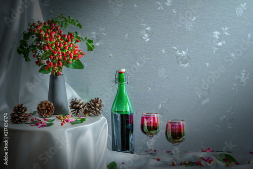 Still life of wine bottle and two winw glasses with a vase of red berry on round table and pine seed on white cover cloth photo