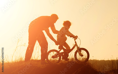 Silhouette of father teaching little daughter to ride a bike at meadow during sunset. Caring father teaches her daughter to ride bicycle