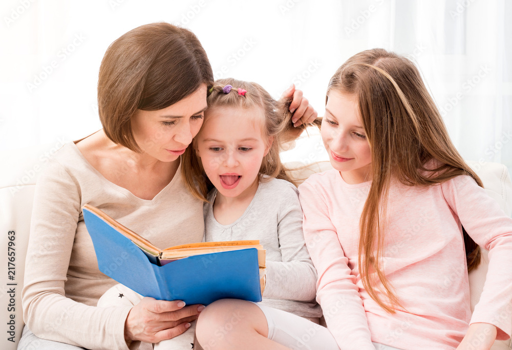 Mom and her two little daughters reading children's book in the living room. Mom and daughters sitting on the coach and reaading Mom and her two little daughters reading children's book in the living room. Mom and daughters sitting on the coach and reaading