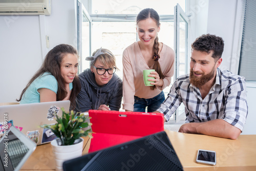 Four co-workers smiling, while watching together a professional business presentation or a funny video on tablet in a modern shared office space for freelancers or young entrepreneurs