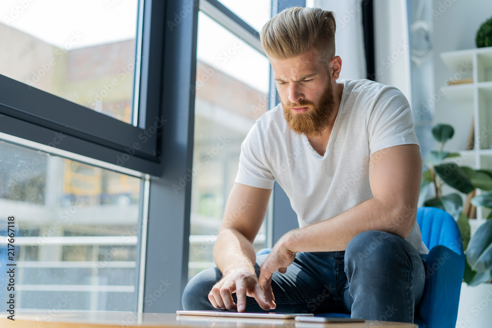 Fototapeta premium Multitasking. Handsome young man wearing glasses and working with touchpad while sitting on the couch in office