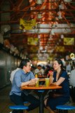 A portrait of an attractive Chinese Asian couple eating at a hawker center. Both are enjoying eating their fried bananas and sugarcane juice.