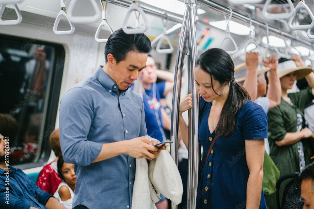 An image of a sweet and young chinese couple in the middle of the train. They are going somewhere to explore. The guy is using his smartphone while the girl is peeking.