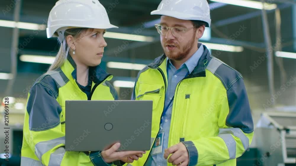 Male and Female Industrial Engineers Wearing safety, Work on a Manufacturing Plant, They Discuss Project while Using Laptop.In the Background Industrial Facility. Male and Female Industrial Engineers Wearing safety, Work on a Manufacturing Plant, They Discuss Project while Using Laptop.In the Background Industrial Facility.