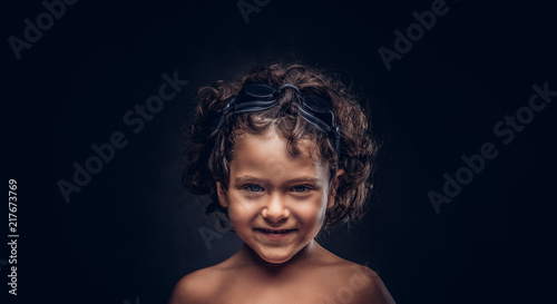 Smiling little shirtless boy in swimming goggles posing in a studio. Isolated on the dark textured background.