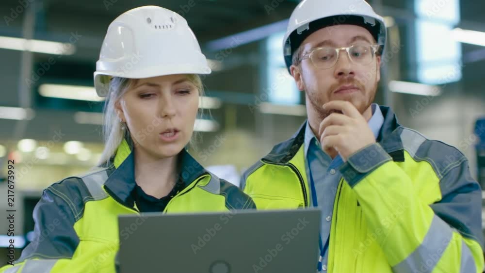 Male and Female Industrial Engineers Work on a Manufacturing Plant, They Discuss Project, Point in the Direction of the Machinery while Using Laptop. Male and Female Industrial Engineers Work on a Manufacturing Plant, They Discuss Project, Point in the Direction of the Machinery while Using Laptop.