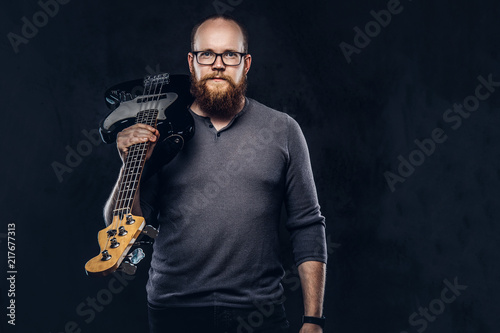Redhead bearded male musician wearing glasses dressed in a gray t-shirt holds electric guitar. Isolated on a dark textured background.