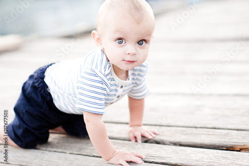 Cute baby boy 1 year old crawling on wooden pier outdoors. Wearing striped clothes. Looking at camera.