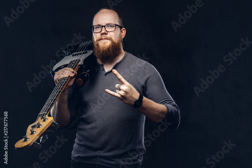 Redhead bearded male musician wearing glasses dressed in a gray t-shirt holds electric guitar and show rock and roll sign. Isolated on a dark textured background.
