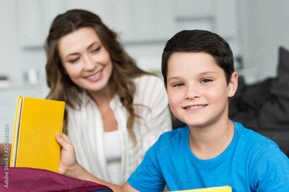 selective focus of smiling boy with book and mother behind at home selective focus of smiling boy with book and mother behind at home