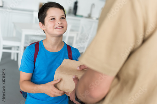 partial view of father giving paper package with food to smiling son with backpack at home