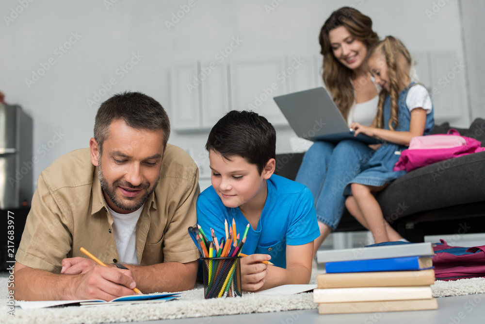 Fototapeta premium selective focus of father and son doing homework together while mother and daughter using laptop on sofa at home