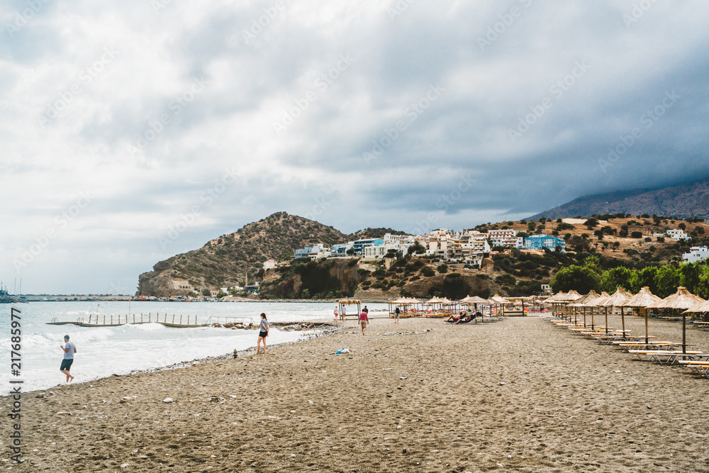Fototapeta premium Crete, Greece. View from cliffs to village with marine vessels, boats and lighthouse. View from cliff on Bay with beach and architecture - vacation destination resort with clear ocean water. Rethymno