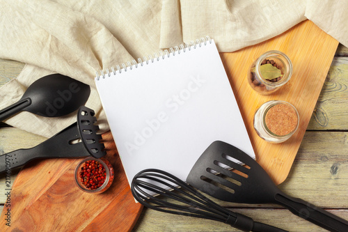 Blank sheet of opened notepad and kitchen utensils on  table with tablecloth, copy space