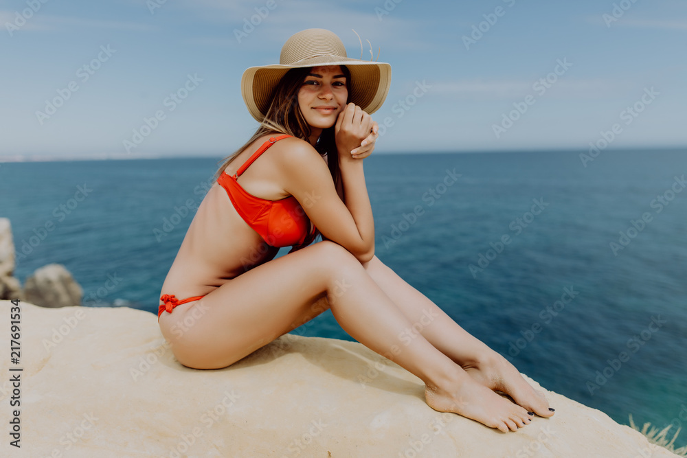 The young woman in red bikini and straw hat sitting on the edge of hill with view on ocean