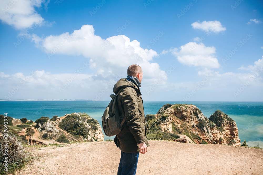 A tourist or traveler with a backpack walks along the coast of the Atlantic Ocean and admires the beautiful view of the ocean near the city called Lagos in Portugal.