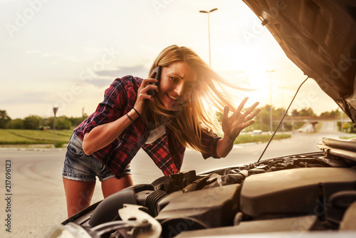 Attractive blonde in front of her car broken down car, assistance concept. Upset woman emotionally reacting on overheated car standing on the roadside with the open hood.