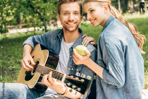 handsome young man playing guitar for his smiling girlfriend and looking at camera while having picnic at park