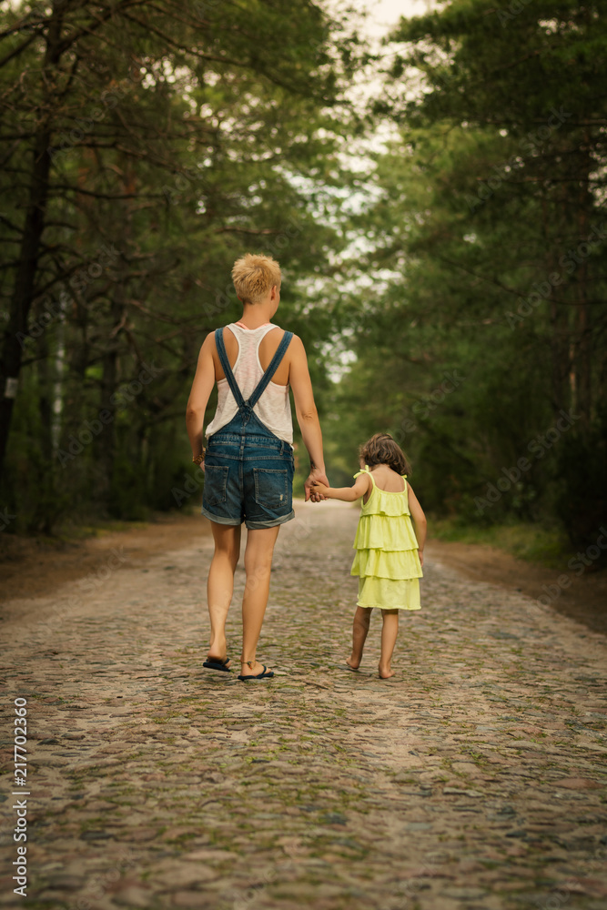 mom and a beautiful daughter in a dress are walking along the road in the summer forest