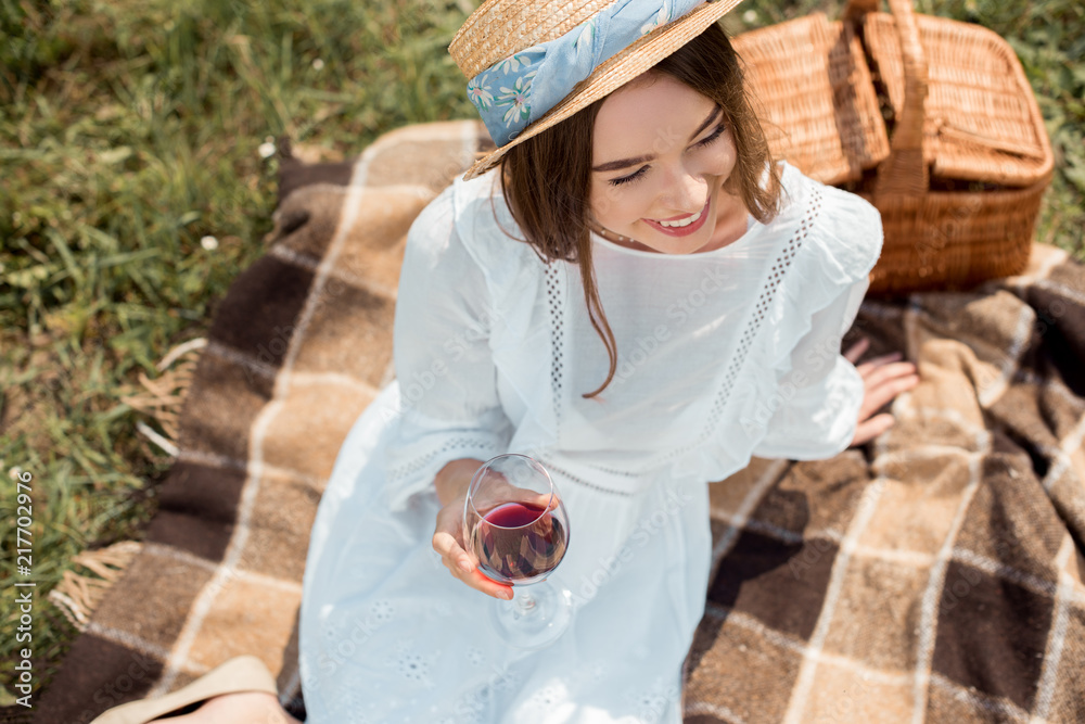 Obraz premium overhead view of smiling woman with glass of red wine resting on blanket in field with wild flowers
