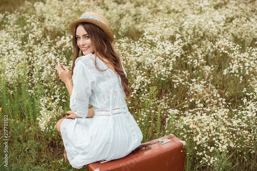 back view of smiling woman in white dress with bouquet of wild camomile flowers sitting on retro suitcase on meadow