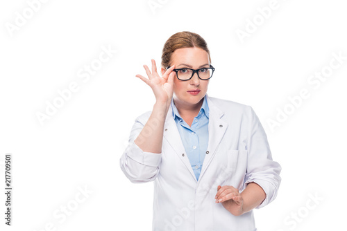 attractive female doctor adjusting eyeglasses and looking at camera isolated on white background