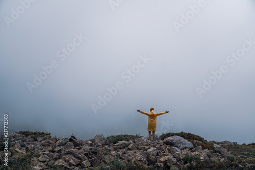 Female in yellow raincoat, jeans shorts standing at top of mountain with view of peaks at horizon. Landscape. Nature.