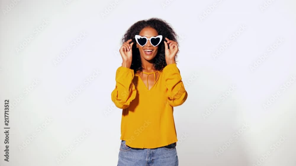 Happy african american woman with afro hair in yellow top wearing heart shaped trendy sunglasses over white wall background. Girl having fun, flirting, smiling to camera. 4k Happy african american woman with afro hair in yellow top wearing heart shaped trendy sunglasses over white wall background. Girl having fun, flirting, smiling to camera. 4k
