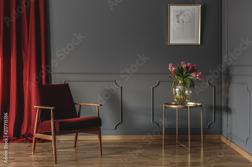 Red curtain and burgundy armchair standing in grey room interior with pink tulips on gold end table and simple poster hanging on the wall with wainscoting