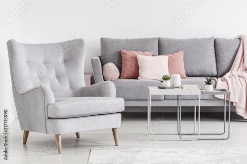 Comfy armchair and grey sofa with pink pillows, and coffee tables in a bright living room interior. Real photo