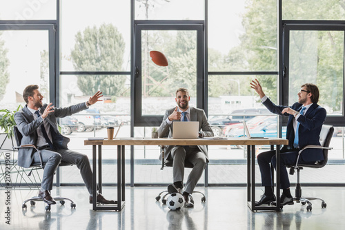 Fototapeta Naklejka Na Ścianę i Meble -  smiling young businessmen using laptops and playing with soccer and rugby balls in office