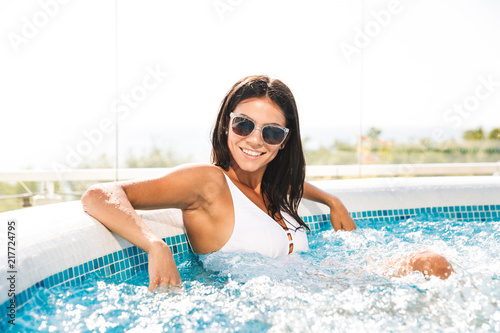 Photo of happy beautiful woman in white swimsuit and sunglasses sitting in jacuzzi hot tub, at luxury hotel zone during vacation