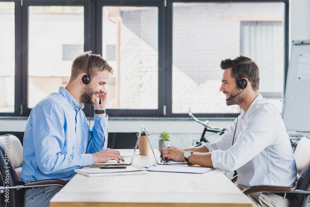 side view of smiling young call center operators in headsets using laptops in office side view of smiling young call center operators in headsets using laptops in office