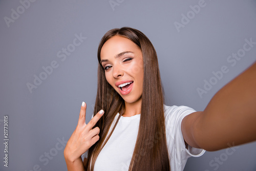 Self-portrait of brown-haired gorgeous attractive nice smiling young lady over grey background, showing v-sign gesture, opened mouth, winking, blinking, flirting, self photographing, isolated