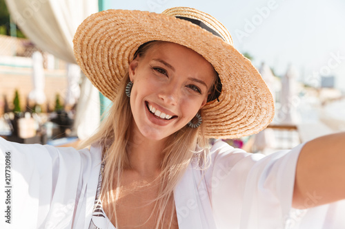 Photo closeup of beautiful young woman 20s in straw hat and swimwear taking selfie, while sunbathing on beach during vacation