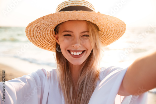 Portrait of caucasian pretty woman 20s in summer straw hat smiling, and taking selfie while walking at seaside