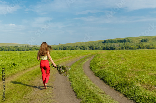 A young girl with a bouquet of flowers moving on a dirt road in the meadow.