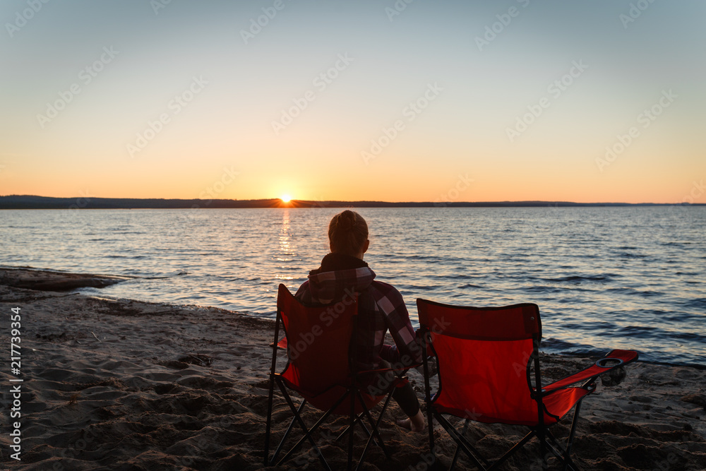 Girl traveler on a folding chair admiring the sunset on the lake. Young woman sitting alone in a folding chair watching the sun setting on the shore at the camping.