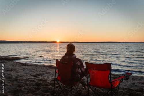 Girl traveler on a folding chair admiring the sunset on the lake. Young woman sitting alone in a folding chair watching the sun setting on the shore at the camping. © korchemkin