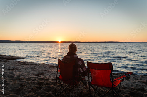 Girl traveler on a folding chair admiring the sunset on the lake. Young woman sitting alone in a folding chair watching the sun setting on the shore at the camping. © korchemkin