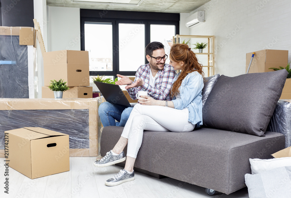 Young couple in new home with notebook sitting on sofa and planning with unpacked boxes in background Young couple in new home with notebook sitting on sofa and planning with unpacked boxes in background
