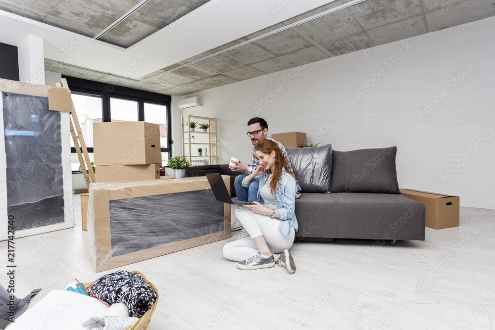 Young couple in new home with notebook sitting on sofa and planning with unpacked boxes in background Young couple in new home with notebook sitting on sofa and planning with unpacked boxes in background