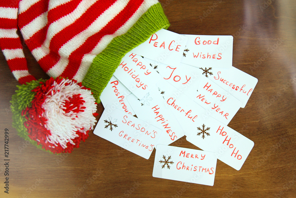 Fototapeta premium Colorful christmas red, white and green knitted Santa claus helper hat, natural wool, handmade knitted, with christmas wishes on paper cards with snowflakes on brown wooden background.