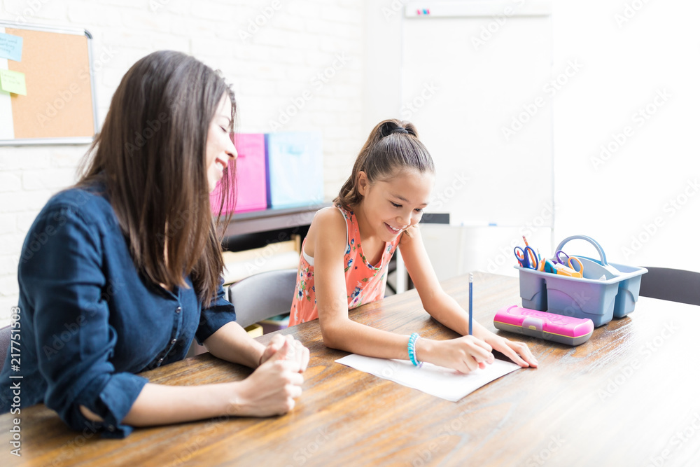 Mother Looking At Daughter Completing School Assignment At Table Mother Looking At Daughter Completing School Assignment At Table