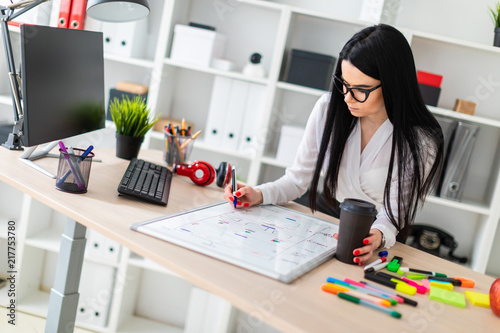 A young girl in glasses stands near a table, holds a glass of coffee in her hand and draws a marker on a magnetic board.