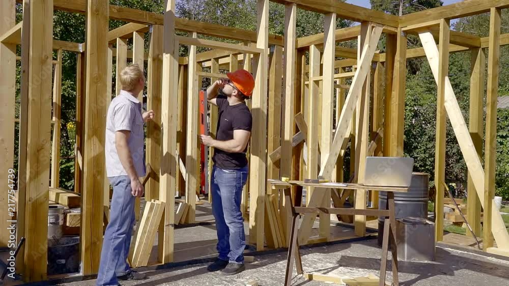 developer and customer are standing inside building house between wooden beams in sunny day, owner is reading papers developer and customer are standing inside building house between wooden beams in sunny day, owner is reading papers